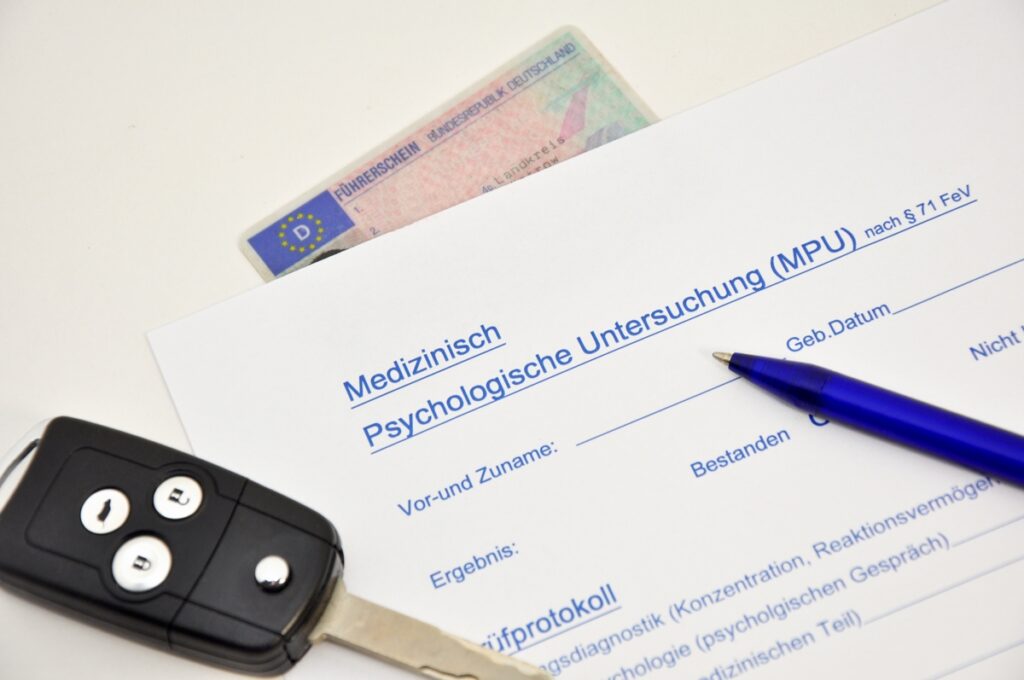 Ein Formular zur MPU, ein Führerschein, ein Autoschlüssel und ein Stift liegen auf einem weißen Tisch.